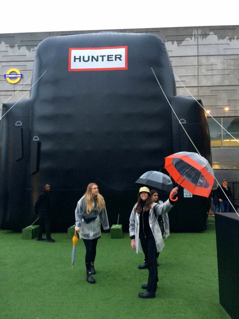 People with Hunter boots and umbrellas in front of giant backpack inflatable